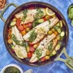 A skillet with cooked white fish fillets showcases a vibrant sea bass recipe, topped with green herb sauce and surrounded by cherry tomatoes, olives, zucchini slices, and lemon wedges. Nearby are a bowl of sauce and fresh basil leaves on a blue tiled surface.