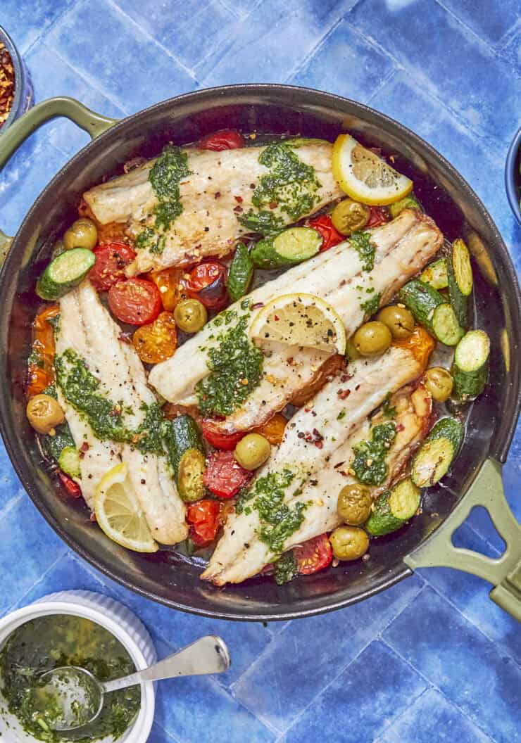 A skillet with cooked white fish fillets showcases a vibrant sea bass recipe, topped with green herb sauce and surrounded by cherry tomatoes, olives, zucchini slices, and lemon wedges. Nearby are a bowl of sauce and fresh basil leaves on a blue tiled surface.