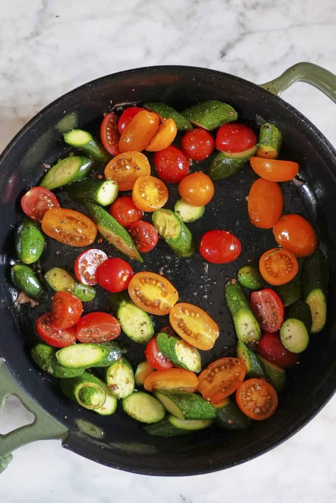 A black skillet with sliced cherry tomatoes and baby cucumbers being sautéed for a sea bass recipe, on a marble countertop. The vibrant red and yellow tomatoes contrast with the green cucumbers.
