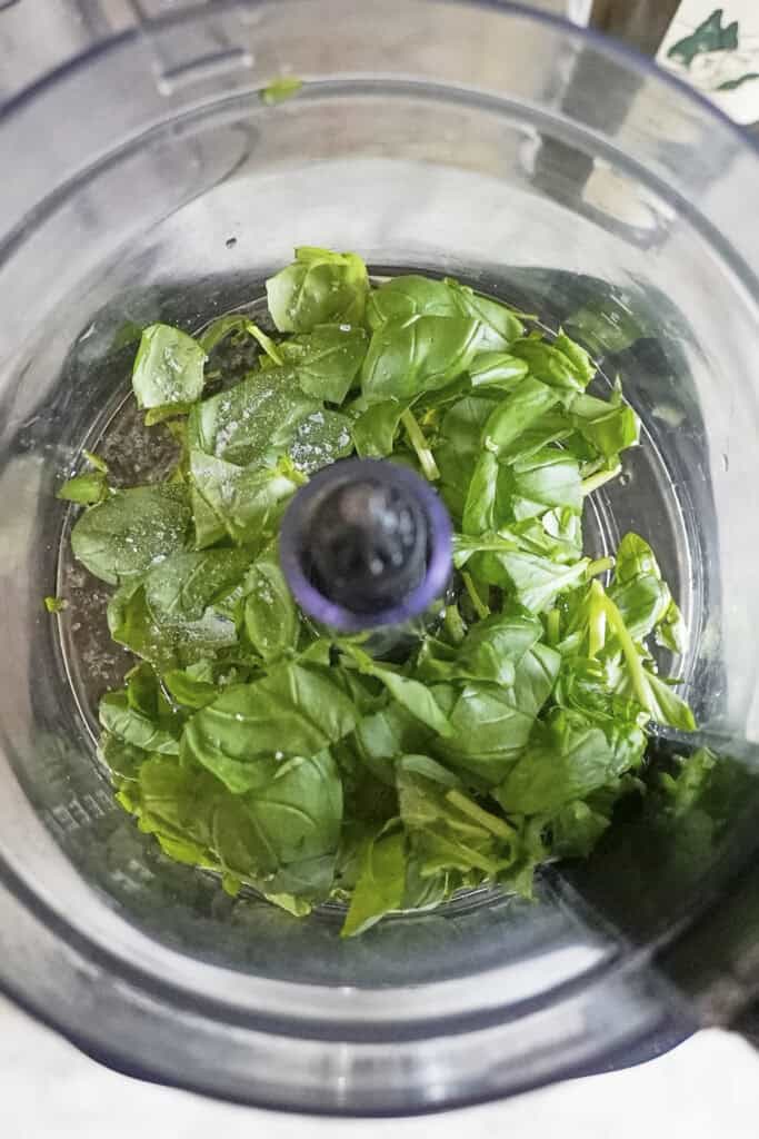 Fresh basil leaves and a sprinkle of salt in the bowl of a food processor, perfect for adding vibrant flavor to your favorite sea bass recipe.
