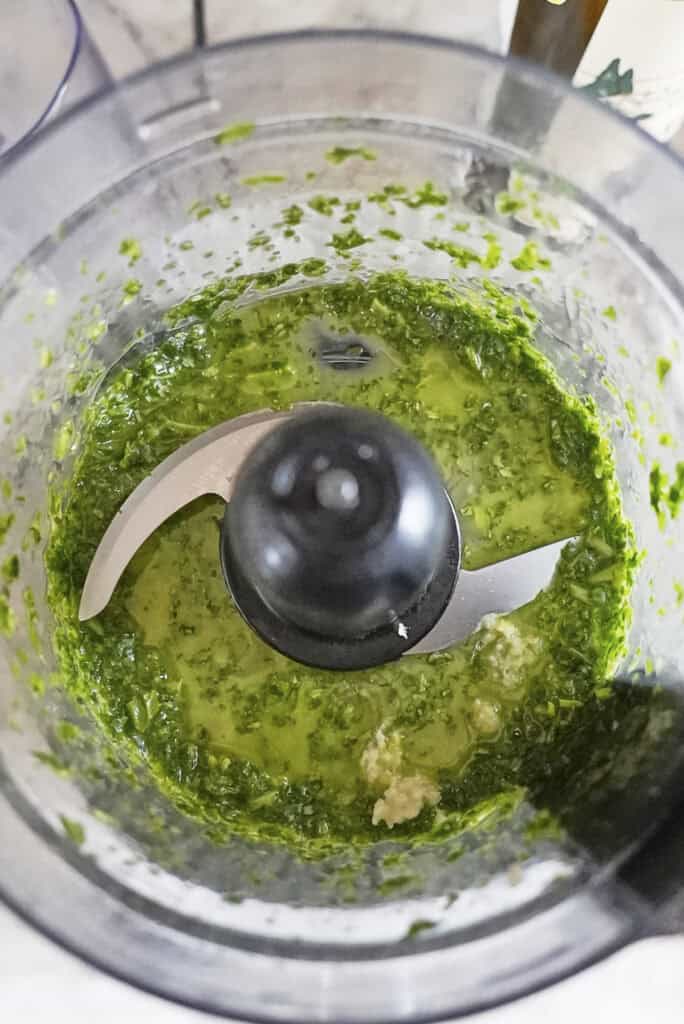 A food processor bowl containing a partially blended green mixture, likely pesto or chimichurri for a sea bass recipe, with visible chopped herbs, oil, and some unblended garlic near the metal blade.