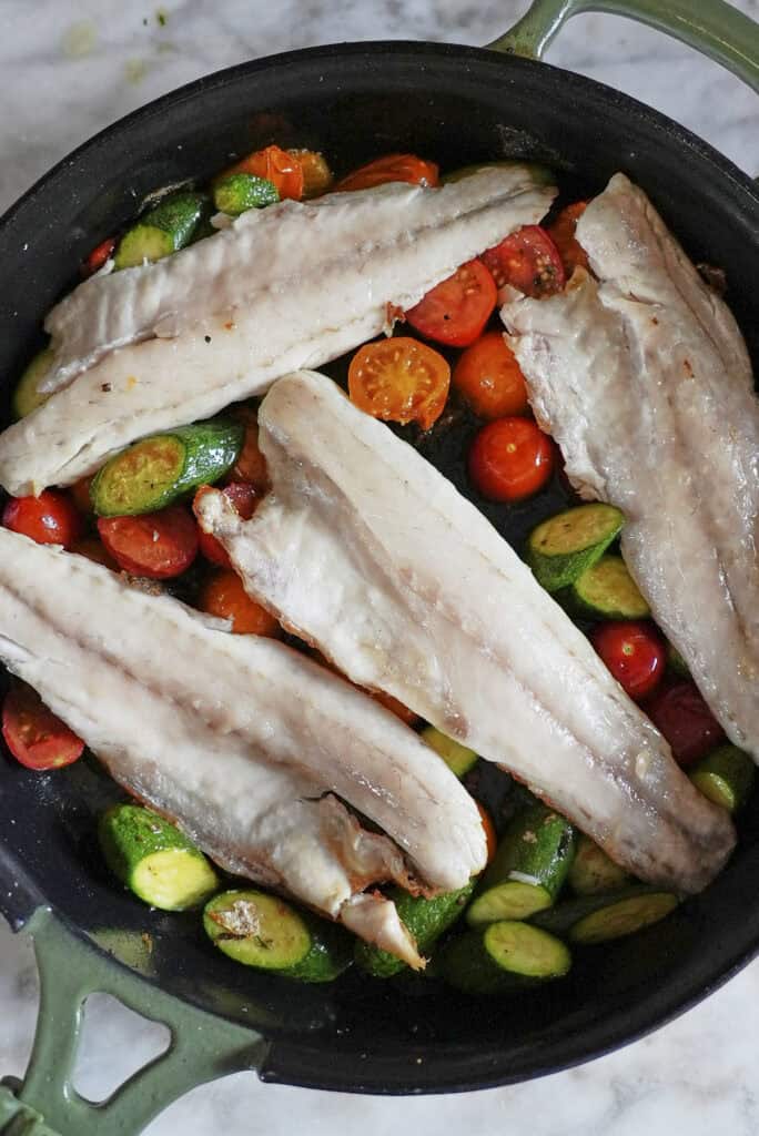 Three raw fish fillets, perfect for a sea bass recipe, are laid over halved cherry tomatoes and sliced zucchini in a black skillet with green handles, ready to be cooked.