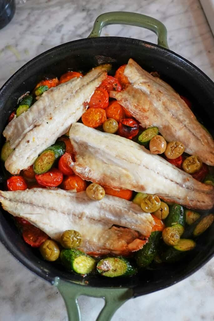 A green-handled pan filled with fillets of white fish, perfect for a sea bass recipe, atop roasted cherry tomatoes, green olives, and sliced zucchini on a marble countertop.