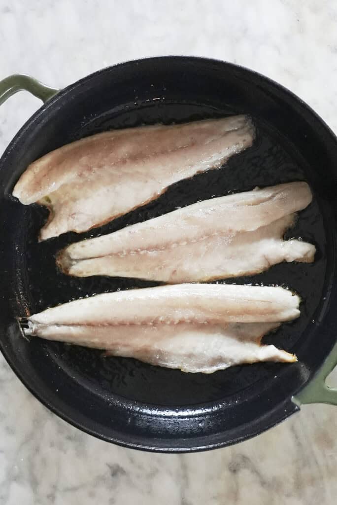 Three raw sea bass fillets are laid side by side in a black pan with oil, ready for your next sea bass recipe. The pan has green handles and rests on a light-colored marble surface.