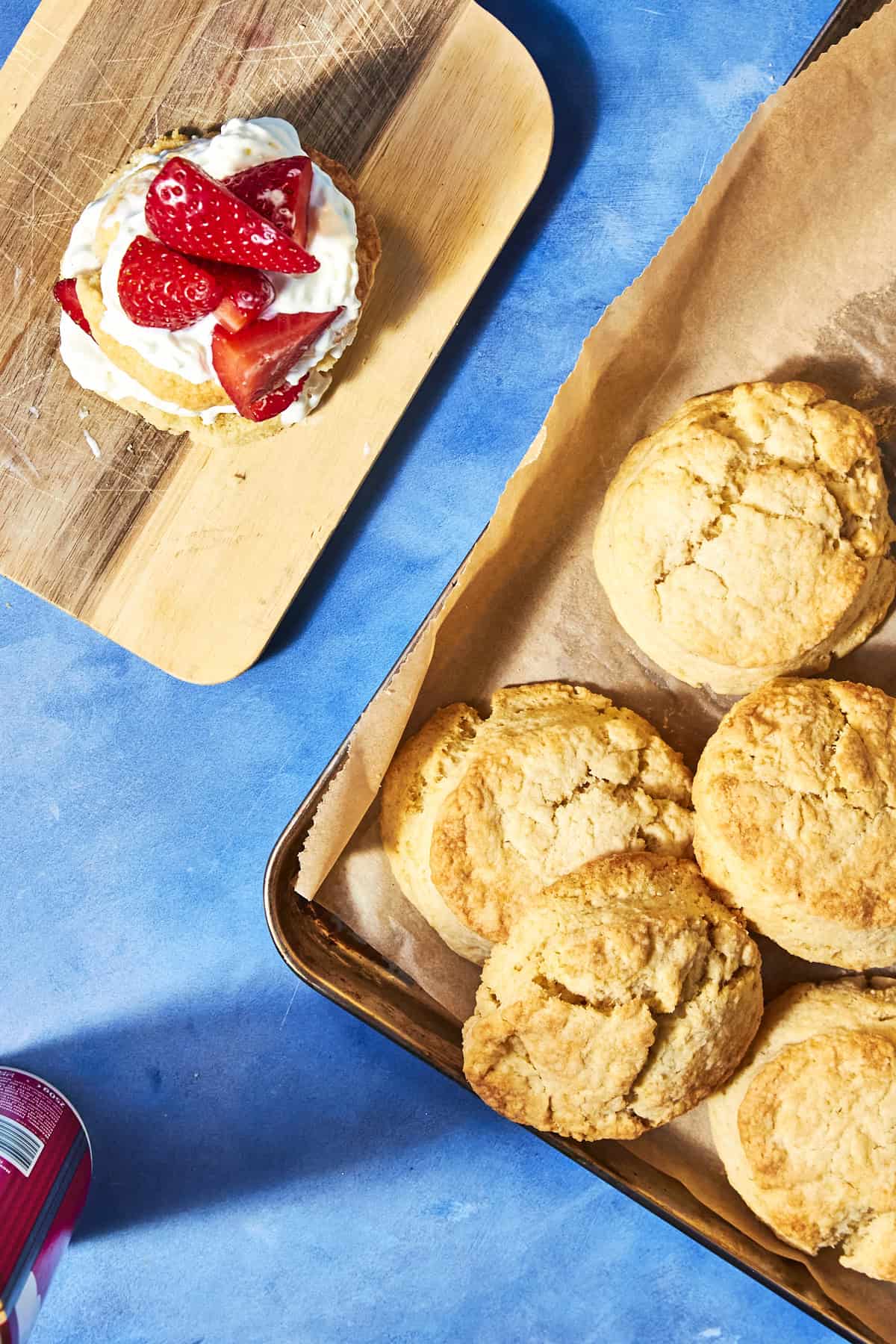 Golden shortcake biscuits on a baking tray with parchment paper, next to a wooden board holding a biscuit topped with whipped cream and fresh sliced strawberries, all on a bright blue surface.