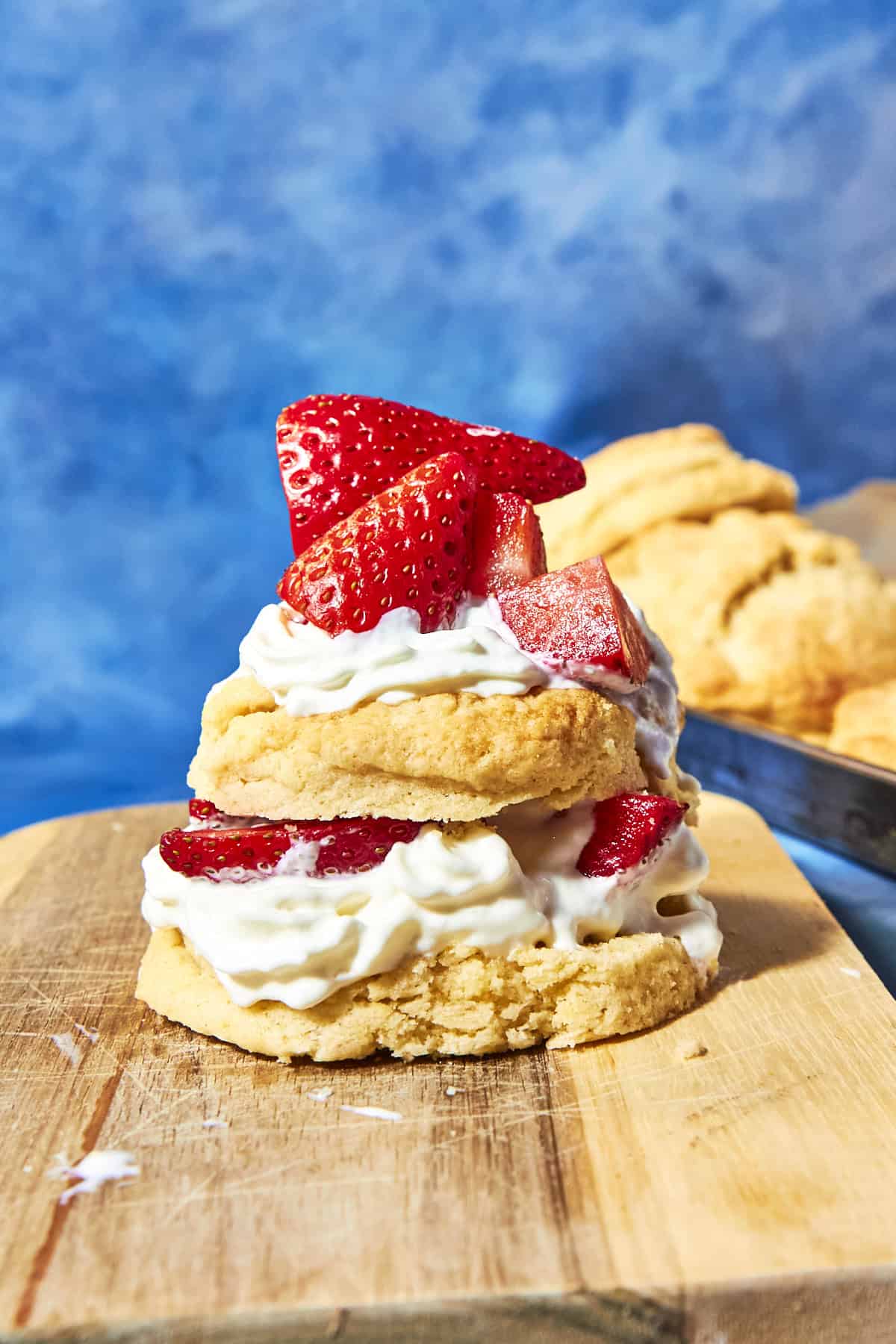 A strawberry shortcake stacked with whipped cream and fresh strawberries sits on a wooden board, showcasing tender shortcake biscuits, with more blurred shortcakes in the background against a blue backdrop.