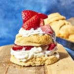 A biscuit split in half and layered with whipped cream and sliced strawberries, forming a strawberry shortcake, sits on a wooden board with extra shortcake biscuits in the blurred background.