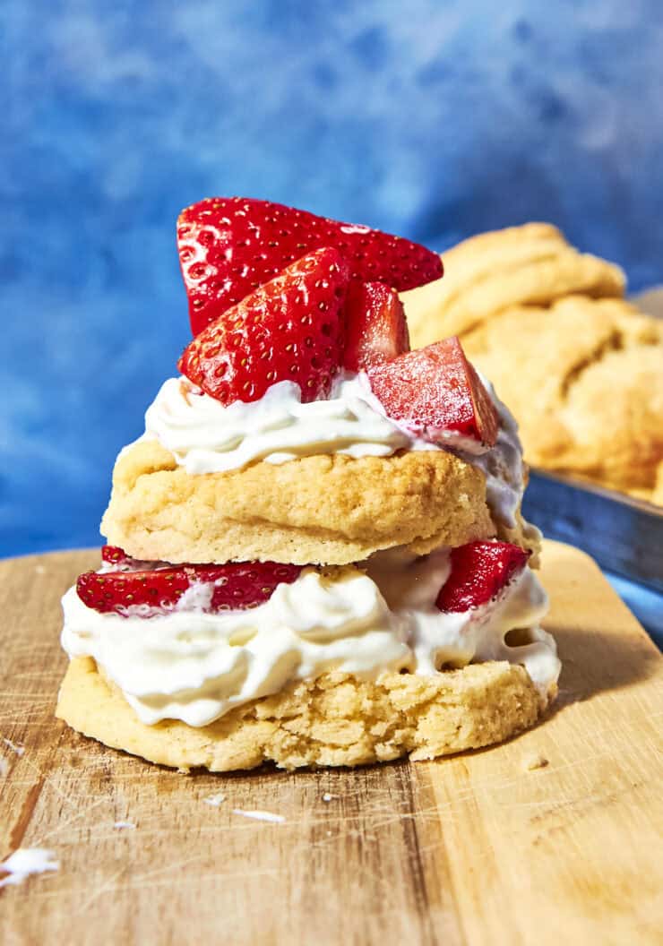 A biscuit split in half and layered with whipped cream and sliced strawberries, forming a strawberry shortcake, sits on a wooden board with extra shortcake biscuits in the blurred background.