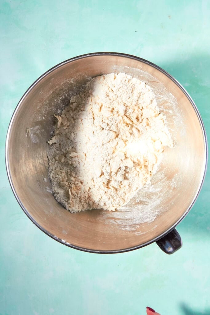 A metal mixing bowl containing partially mixed, crumbly shortcake biscuit dough sits on a light turquoise surface.