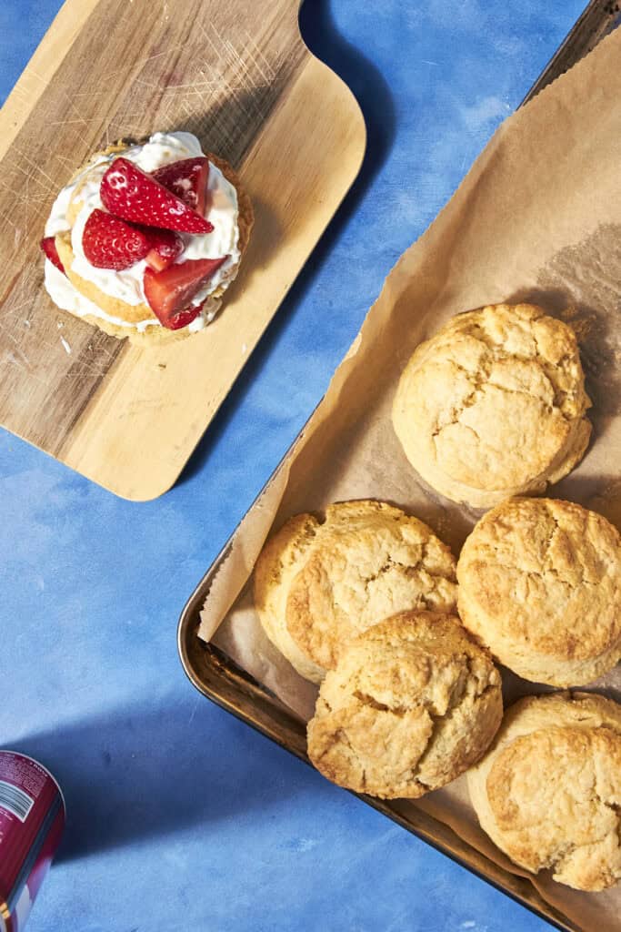 Golden shortcake biscuits rest on a parchment-lined tray, while a wooden board displays a biscuit topped with whipped cream and fresh strawberries. The scene is set on a blue surface.