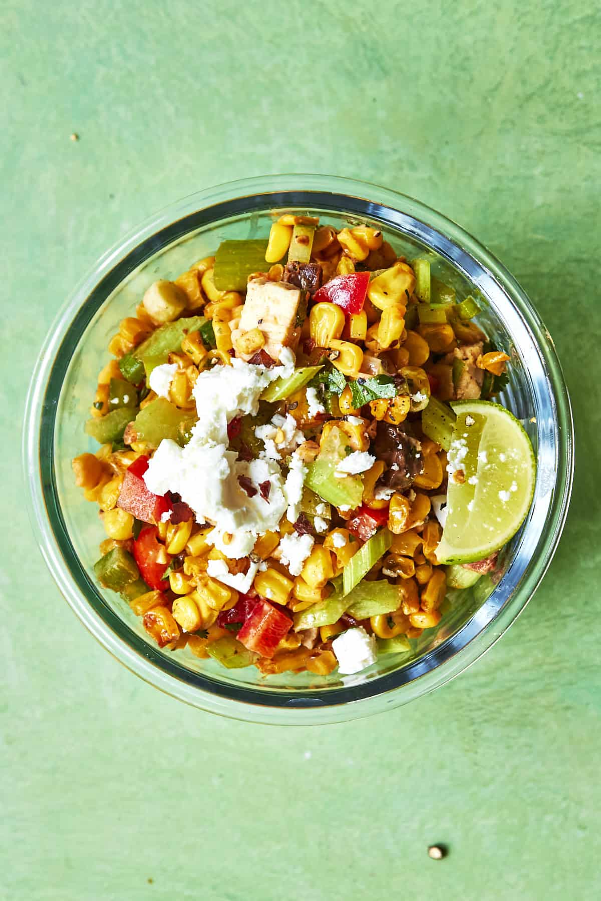 A glass bowl filled with a colorful roasted corn salad, featuring grilled corn, diced red and green peppers, crumbled white cheese, and a lime wedge, set on a light green surface.