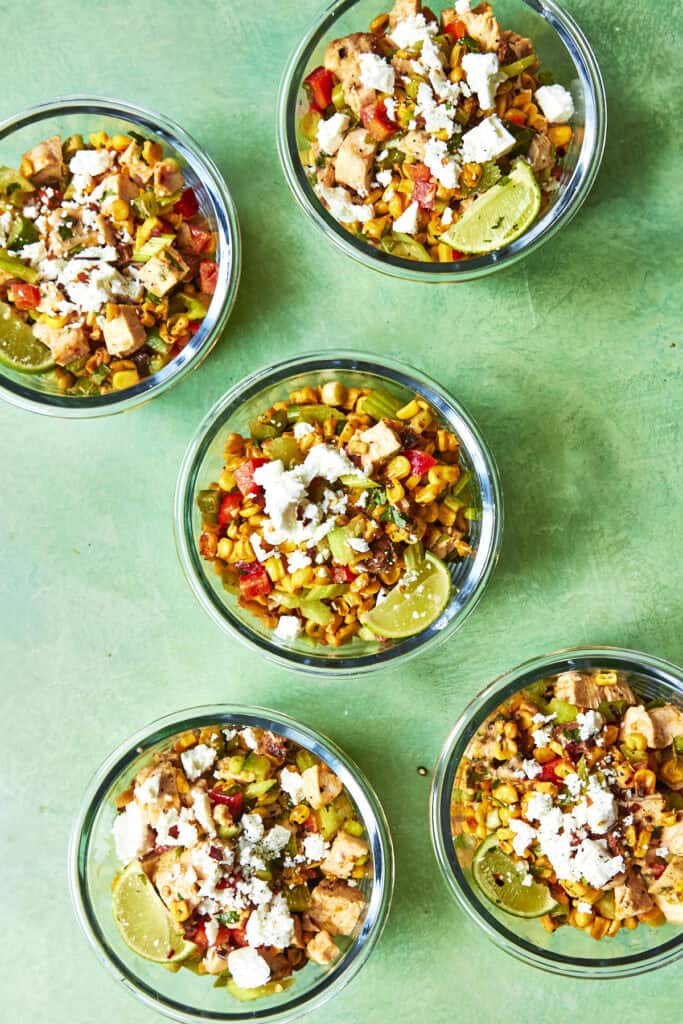 Five glass bowls filled with colorful Mexican-style rice salad, topped with crumbled cheese, diced chicken, roasted corn, mixed vegetables, and lime wedges, arranged on a green surface.