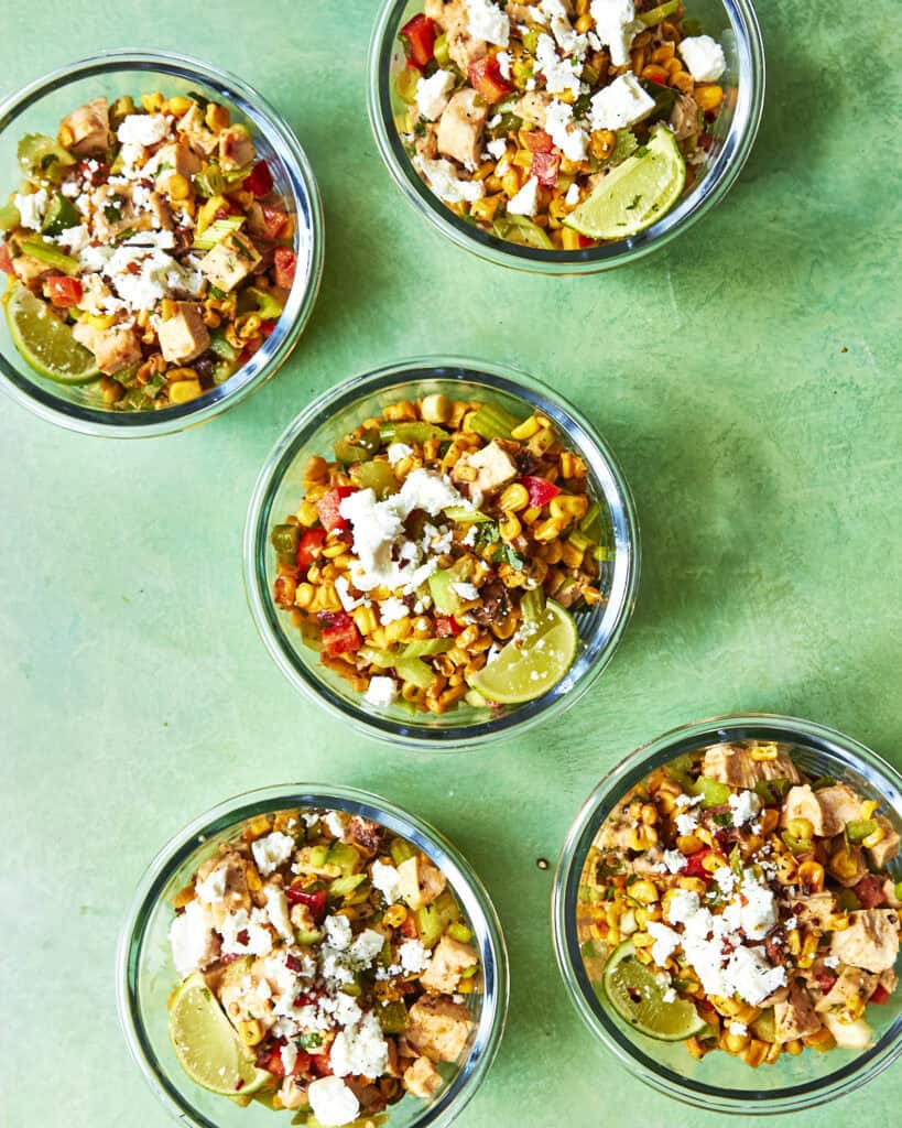 Five glass bowls filled with colorful Mexican street corn salad made with roasted corn, garnished with lime wedges and crumbled cheese, are arranged on a green surface. The salad contains corn, diced vegetables, and small pieces of protein.
