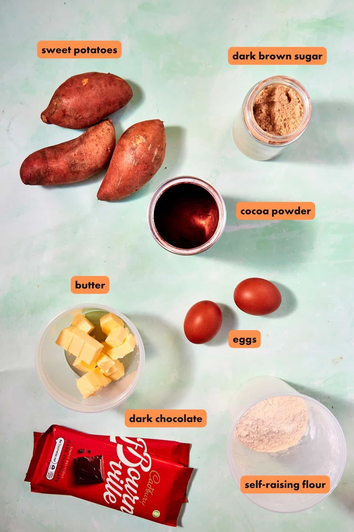 Flat lay of ingredients on a light surface: three sweet potatoes, dark brown sugar, cocoa powder, butter cubes, eggs, a red chocolate bar, self-raising flour—and labeled for easy use. Ideal for baking Funfetti Cookies or experimenting with new treats.