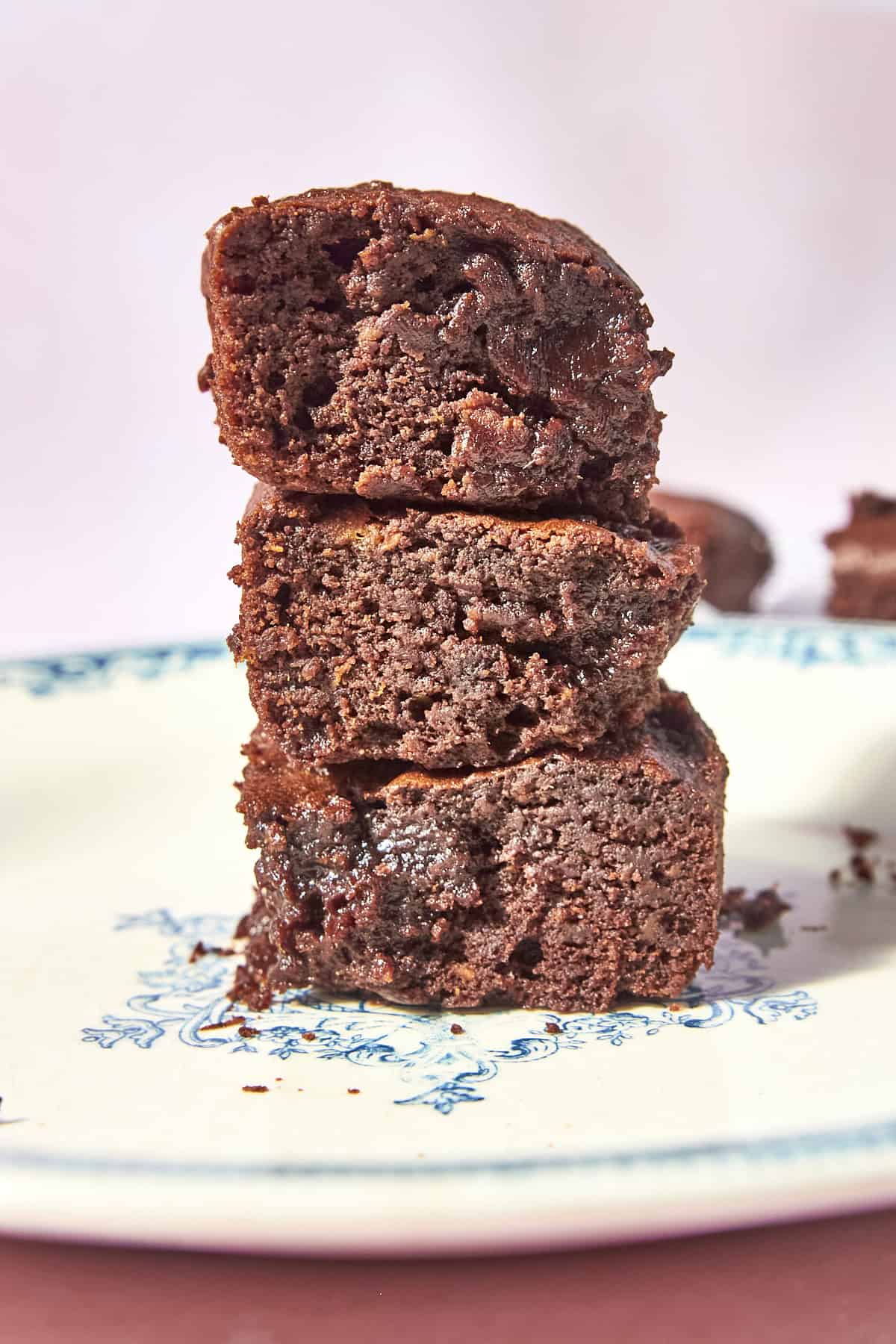 A stack of three gooey chocolate brownies is placed on a white plate with a blue floral pattern, with one brownie showing a melted chocolate center.