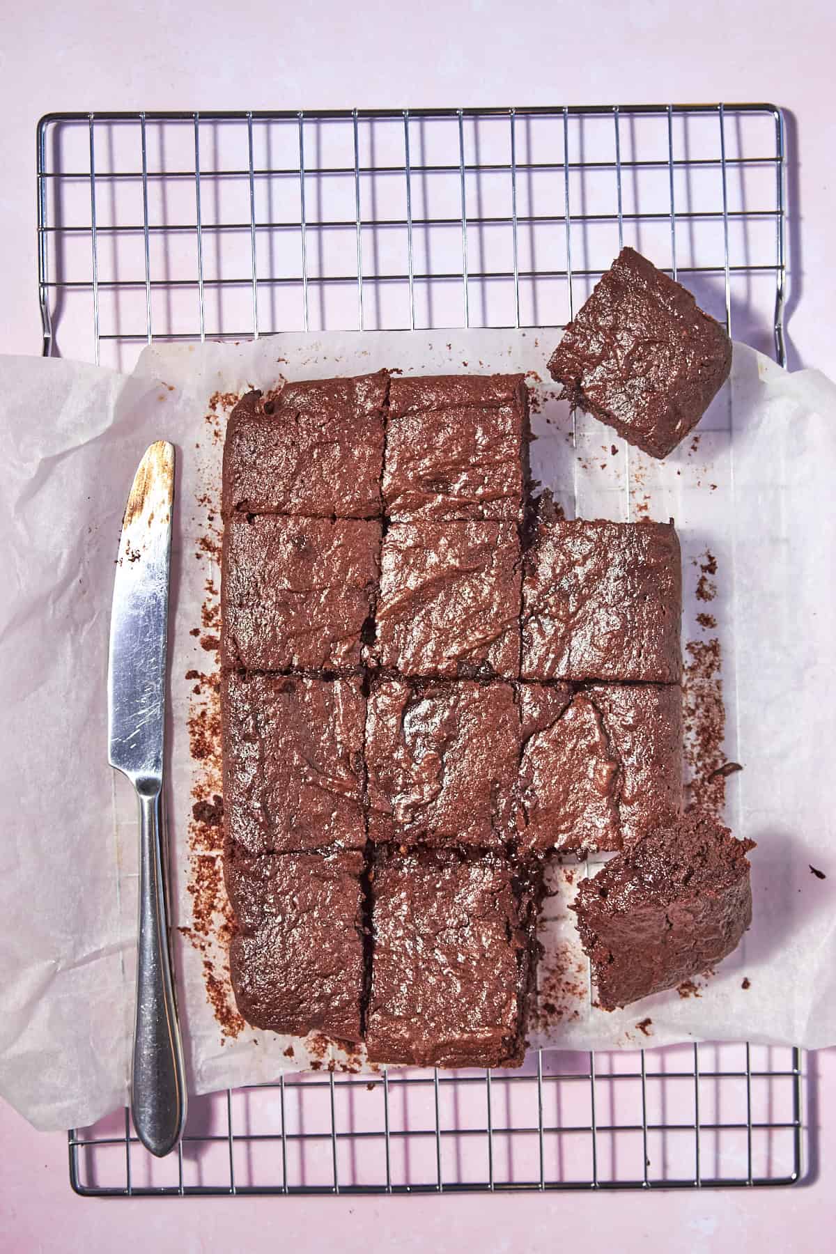 Nine chocolate brownies on parchment paper, with two pieces separated, sit on a cooling rack. A butter knife with chocolate crumbs rests beside the brownies on a light pink background.