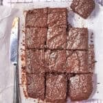 A batch of chocolate brownies cut into squares, with one piece slightly separated, sits on parchment paper atop a cooling rack. A butter knife with chocolate smears lies to the left of the brownies.