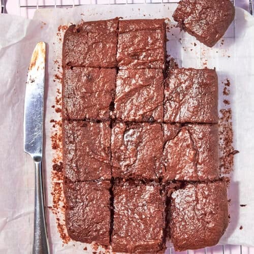 A batch of chocolate brownies cut into squares, with one piece slightly separated, sits on parchment paper atop a cooling rack. A butter knife with chocolate smears lies to the left of the brownies.