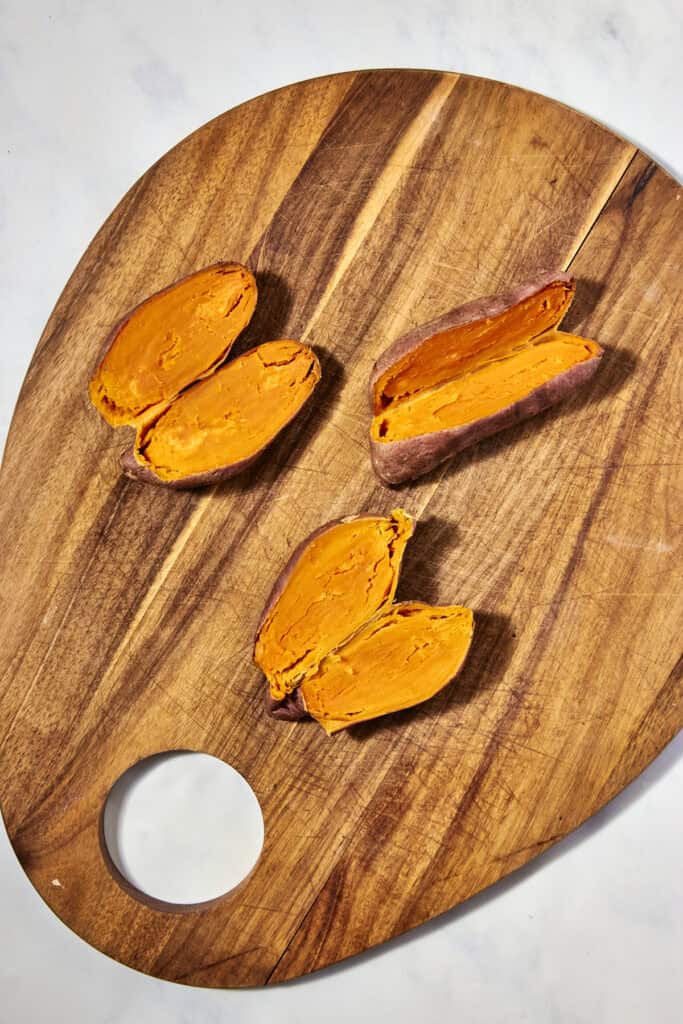 Three cooked sweet potatoes, cut in half to reveal their orange flesh, are arranged on a wooden cutting board with a circular hole, set on a white marble surface.