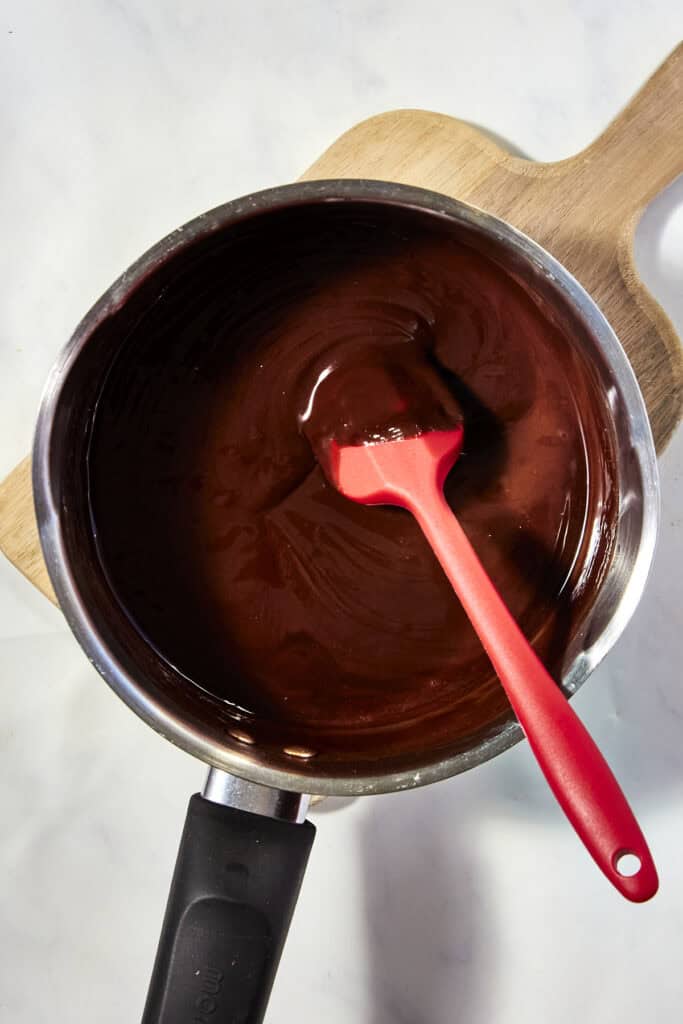 A saucepan filled with melted chocolate being stirred by a red spatula, placed on a wooden board against a light background.