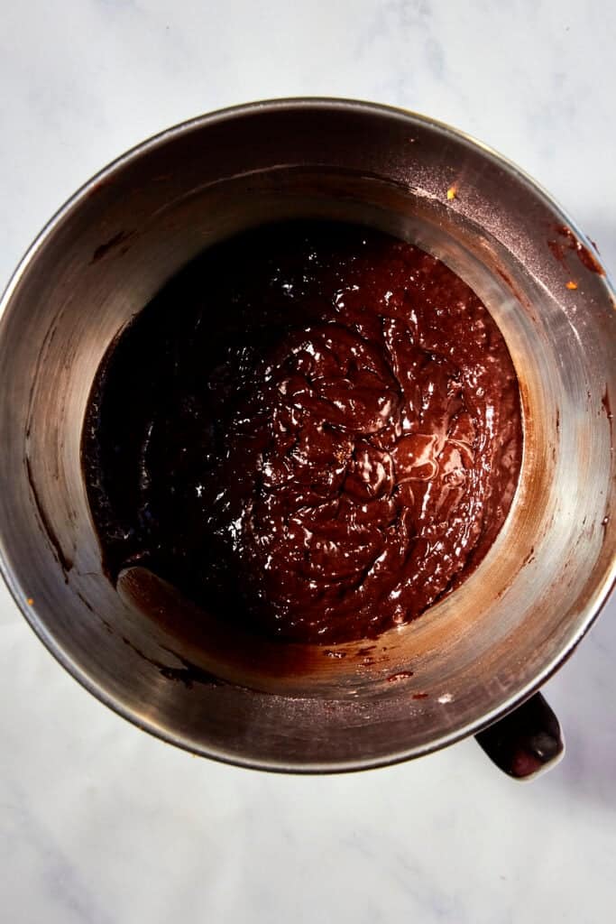 A metal mixing bowl filled with thick, glossy chocolate batter sits on a white marble surface.