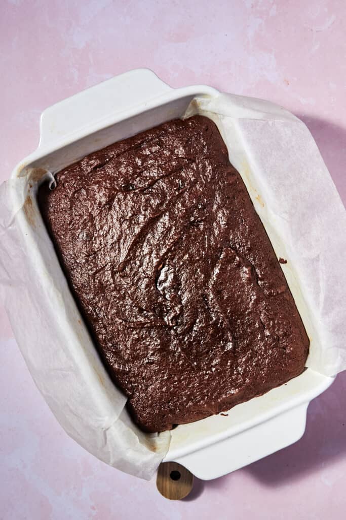 A batch of chocolate brownies in a white baking dish lined with parchment paper, resting on a pink surface.