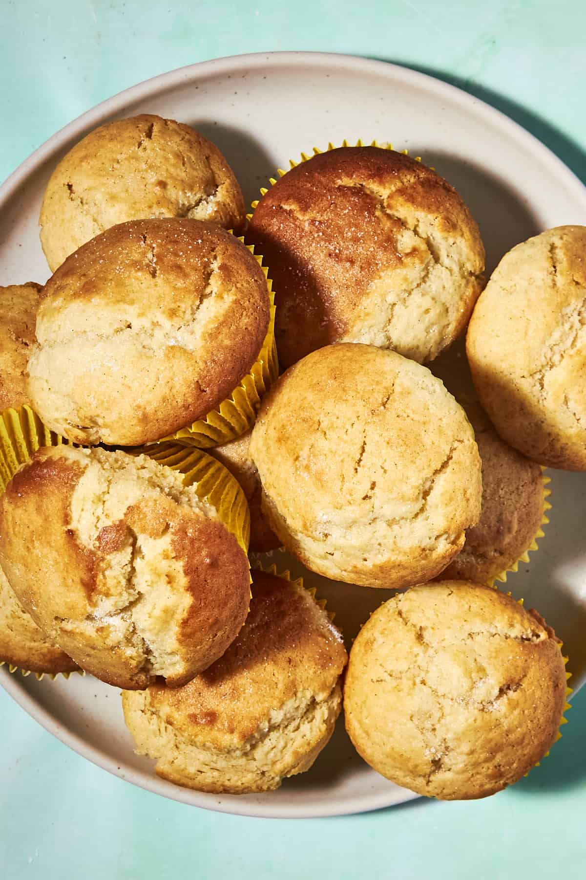 A plate filled with golden brown muffins in yellow paper liners sits on a light blue surface. The muffins appear fluffy and freshly baked.