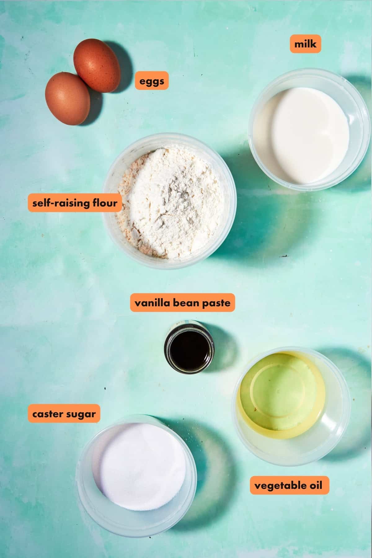 Flat lay of baking ingredients on a teal background for Funfetti Cookies: two eggs, self-raising flour, caster sugar, milk, vegetable oil, and vanilla bean paste, each clearly labeled with orange text.