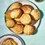 A plate of golden-brown muffins, some in yellow wrappers, sits on a light blue surface. Nearby, a blue and white plate holds a halved muffin, and a small jar with a spoon is in the corner.