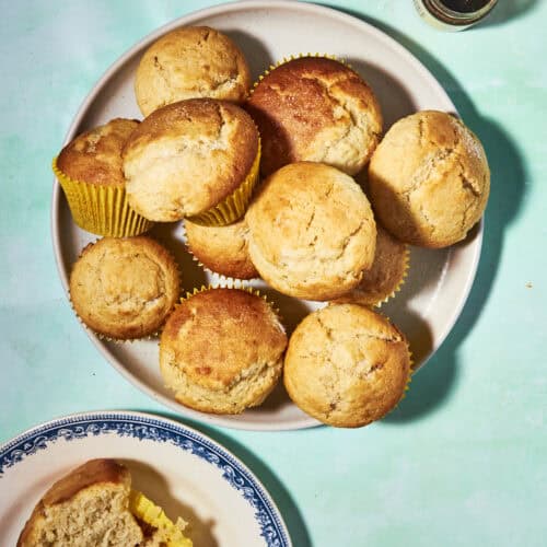 A plate of golden-brown muffins, some in yellow wrappers, sits on a light blue surface. Nearby, a blue and white plate holds a halved muffin, and a small jar with a spoon is in the corner.