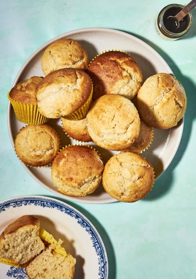 A plate of golden-brown muffins, some in yellow wrappers, sits on a light blue surface. Nearby, a blue and white plate holds a halved muffin, and a small jar with a spoon is in the corner.