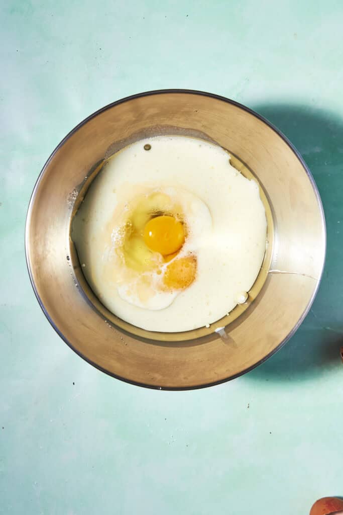A metal mixing bowl containing milk and a raw egg on a light blue surface, viewed from above.