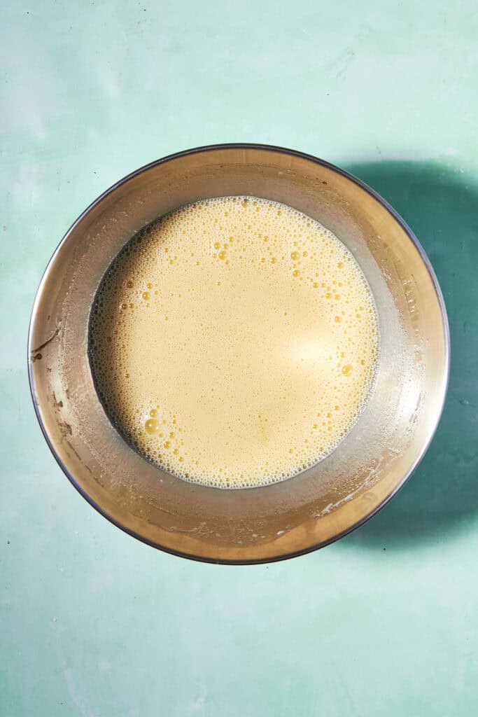 A stainless steel mixing bowl filled with a pale, frothy liquid, likely whisked eggs or batter, sits on a light teal surface.