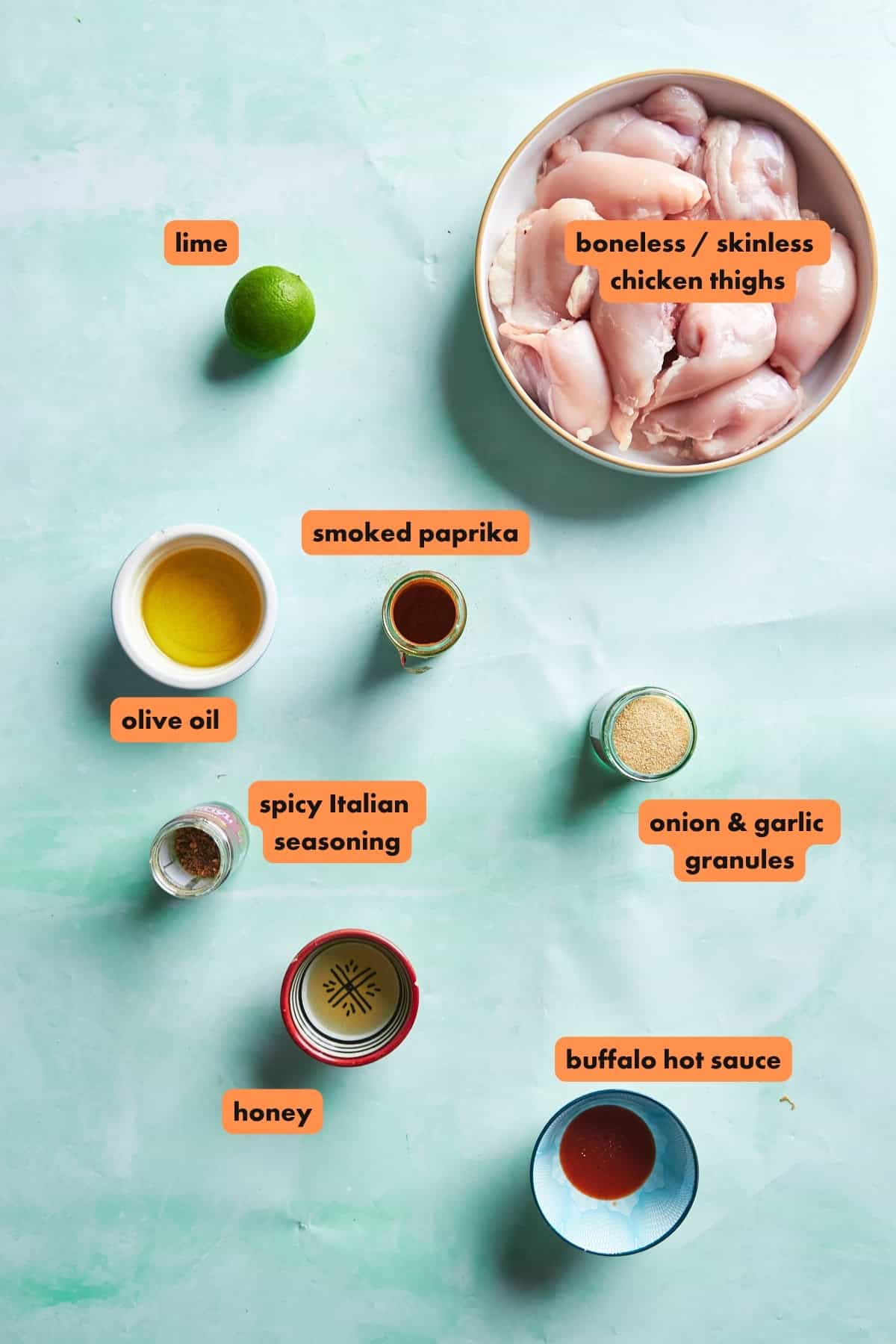 A flat lay photo of ingredients for a chicken recipe: a bowl of boneless chicken thighs, a whole lime, olive oil, smoked paprika, spicy Italian seasoning, honey, onion & garlic granules, and buffalo hot sauce.