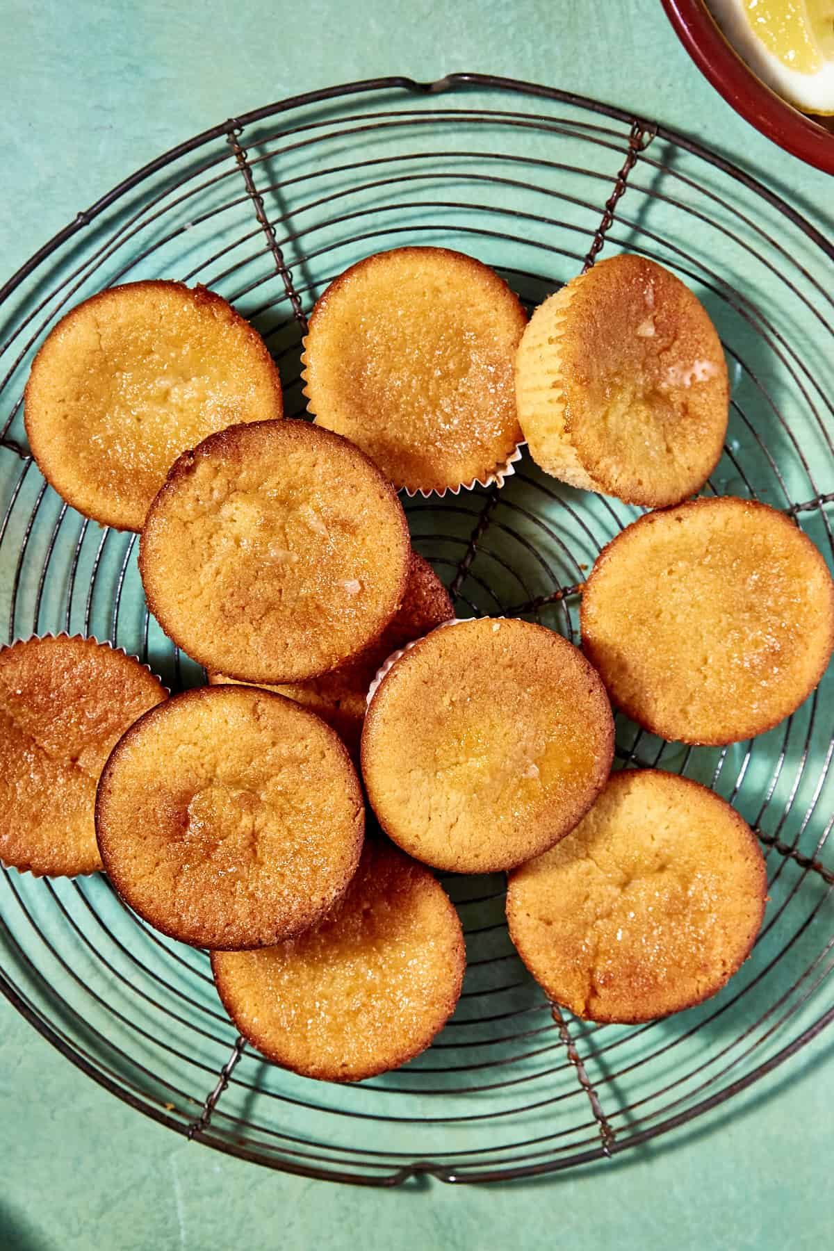 Golden brown lemon muffins arranged on a round wire cooling rack, set on a teal surface. One muffin has a visible paper liner, and the muffins have slightly crisp, caramelized tops.