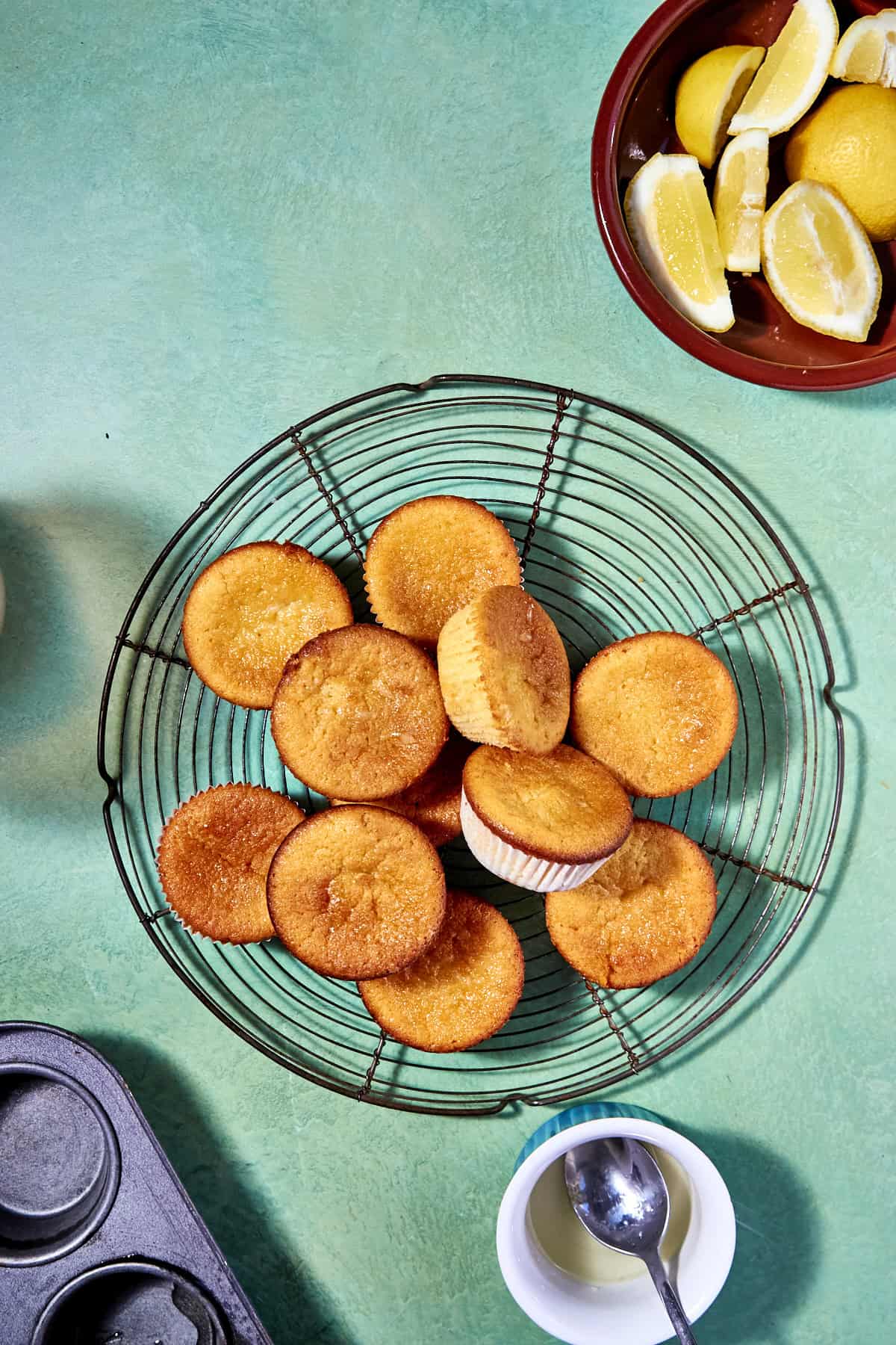 A wire rack holds several golden-brown lemon muffins on a turquoise surface, with a bowl of lemon wedges, an empty muffin tin, and a small bowl of white sauce nearby.