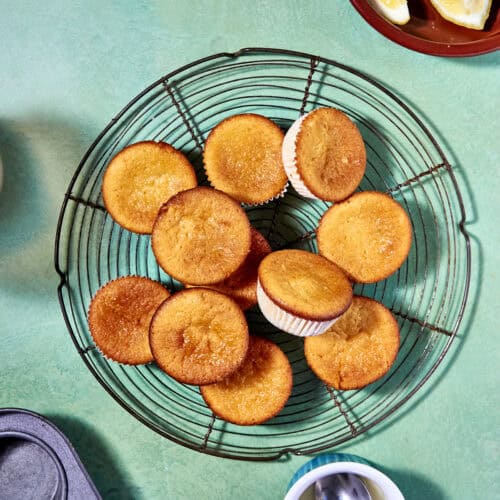 A wire cooling rack holds a pile of golden brown lemon muffins; nearby are a muffin tin, a bowl of lemon wedges, and a small bowl of syrup, all on a green surface.