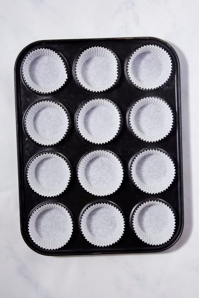 A black muffin tin with twelve cups, each lined with a white paper cupcake liner ready for lemon muffins, photographed from above on a light surface.