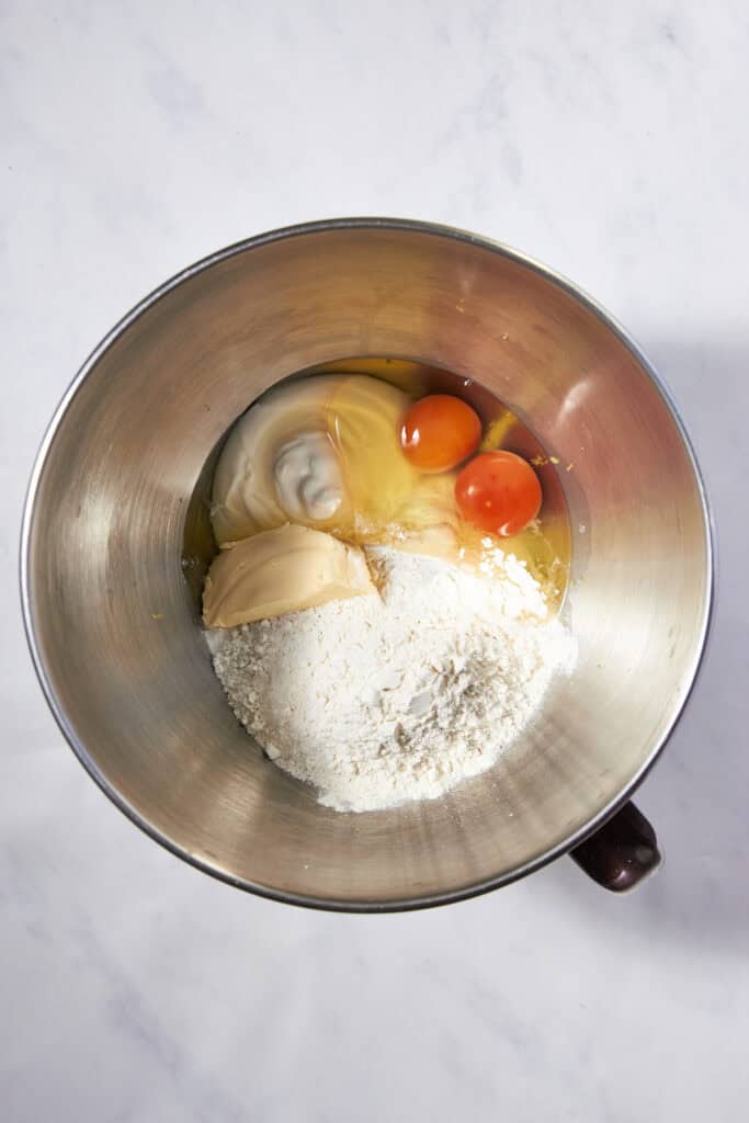 A mixing bowl containing flour, two eggs, a dollop of peanut butter, and a scoop of yogurt or sour cream sits on a white marble surface—perfect for whipping up delicious lemon muffins.