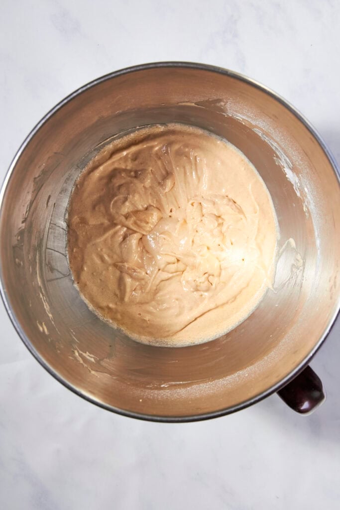A metal mixing bowl with a light brown, creamy batter mixture for lemon muffins sits on a white countertop.