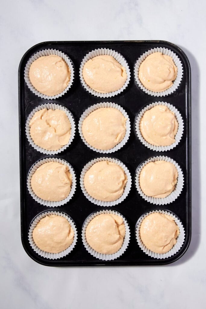 A black muffin tray filled with twelve paper cupcake liners, each containing unbaked lemon muffins batter, set on a white marble surface.