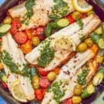 A pan with cooked sea bass fillets is topped with green herb sauce and surrounded by cherry tomatoes, olives, and lemon slices on a blue patterned surface. Text reads Best Sea Bass Recipe and www.beatthebudget.com—a must-try sea bass recipe.
