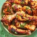 A bowl of baked chicken drumsticks garnished with chopped herbs sits on a green surface, with text reading Oven-Baked Chicken Drumsticks and Fail Proof Ultimate Method.