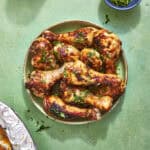 A plate of grilled chicken drumsticks garnished with chopped herbs sits on a green surface, with a small bowl of more herbs nearby. Part of a foil-covered dish is visible in the corner.