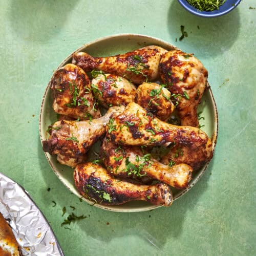 A plate of grilled chicken drumsticks garnished with chopped herbs sits on a green surface, with a small bowl of more herbs nearby. Part of a foil-covered dish is visible in the corner.