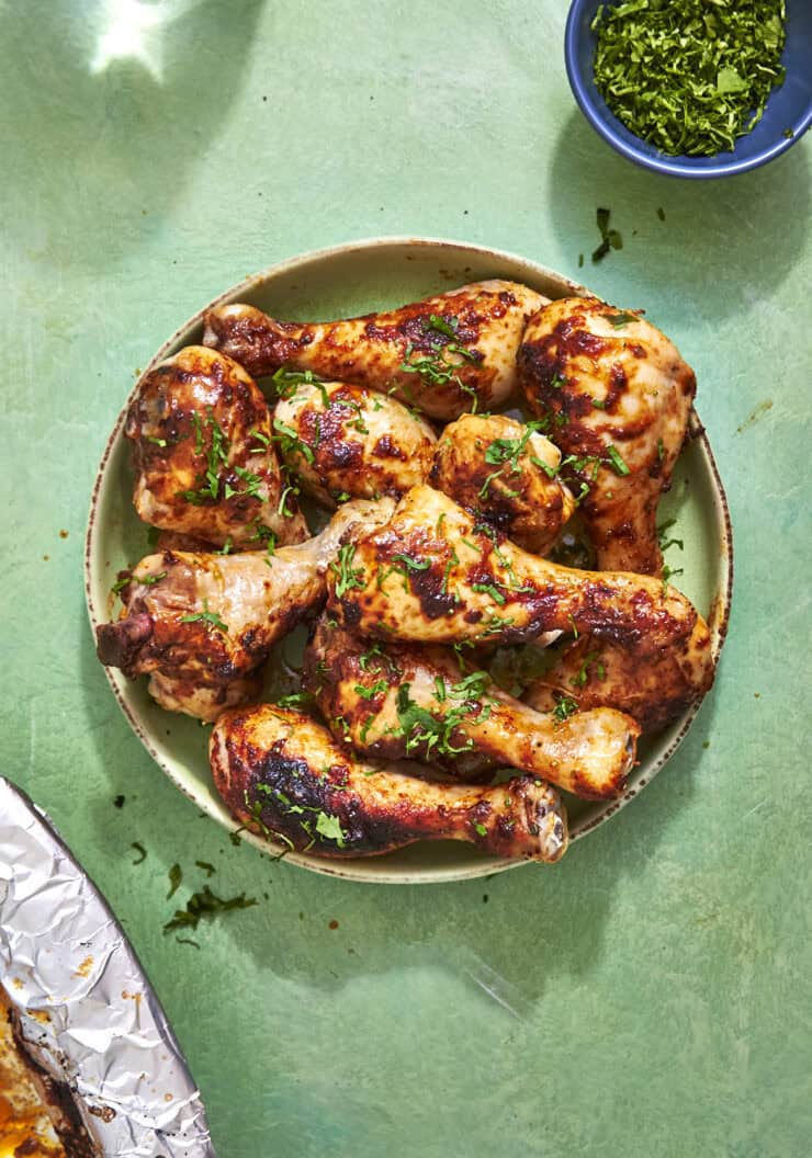 A plate of grilled chicken drumsticks garnished with chopped herbs sits on a green surface, with a small bowl of more herbs nearby. Part of a foil-covered dish is visible in the corner.