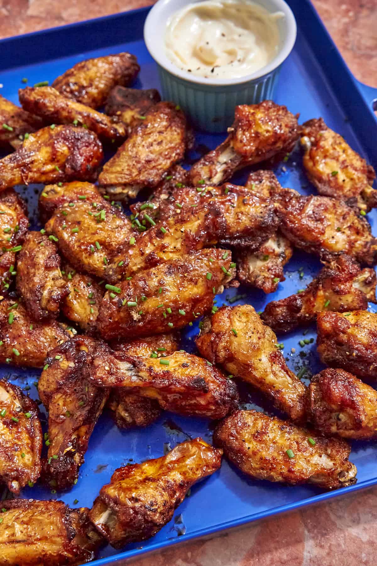 A blue tray filled with flavorful baked chicken wings, garnished with chopped herbs and served alongside a small dish of creamy dipping sauce in the corner.