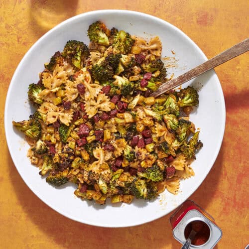 A white plate filled with bow-tie pasta, roasted broccoli, diced sausage, and yellow vegetables sits on an orange surface. A wooden spoon rests on the plate. Nearby are an open spice tin and a glass jar of red pepper flakes.