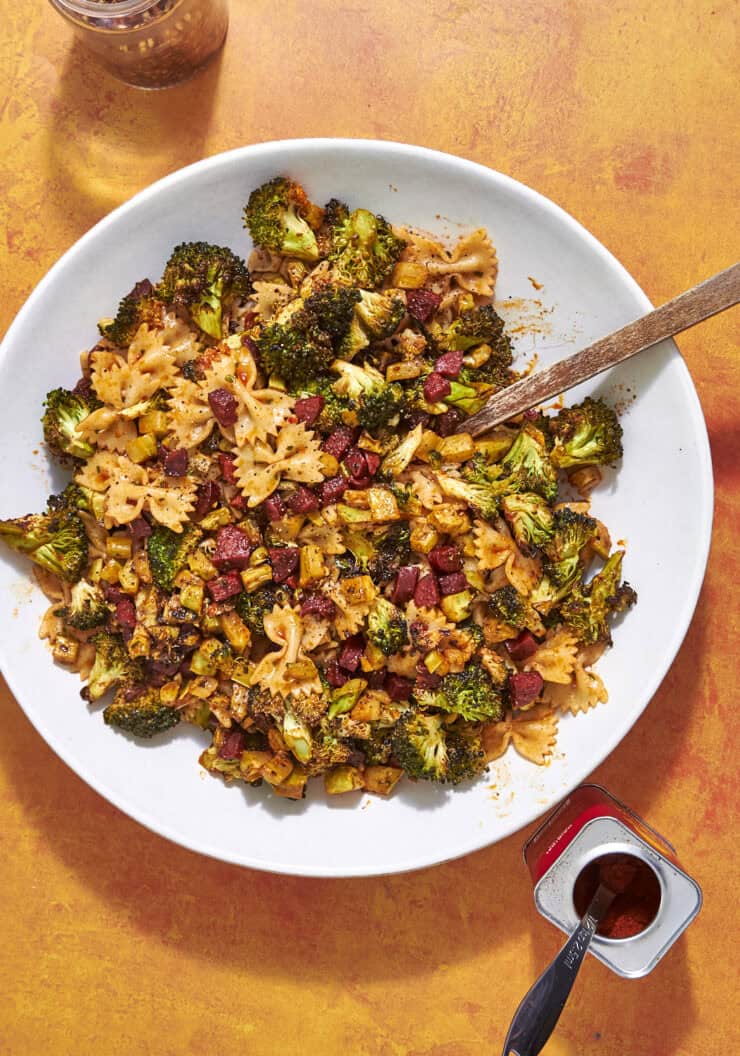 A white plate filled with bow-tie pasta, roasted broccoli, diced sausage, and yellow vegetables sits on an orange surface. A wooden spoon rests on the plate. Nearby are an open spice tin and a glass jar of red pepper flakes.