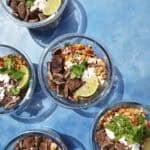 Five glass bowls containing beef strips, roasted corn, crumbled cheese, cilantro, and lime wedges are arranged on a bright blue surface with strong sunlight casting shadows.